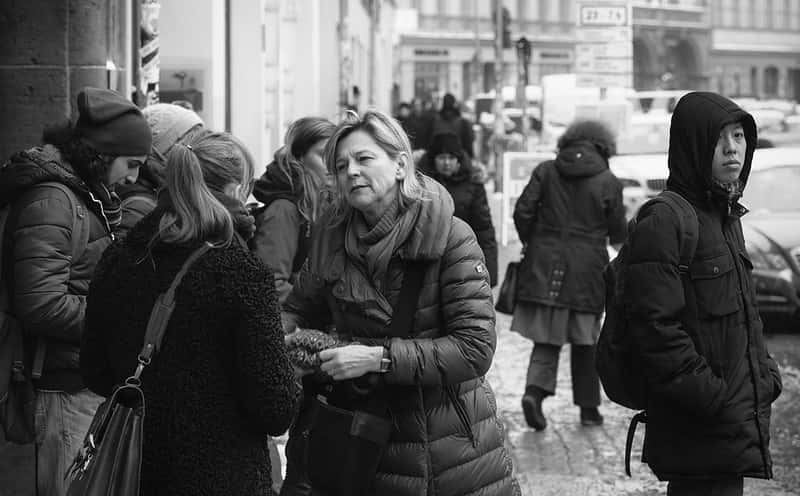 Menschen die sich unterhalten in einer Winterszene in der Stadt. Ein Mann (Asiate) schaut in die Kamera, steht aber abseits der Szene.