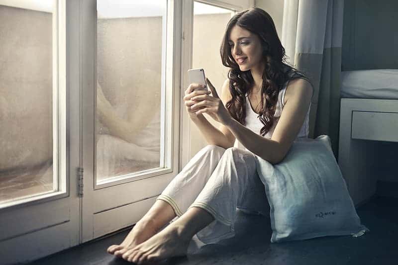 a-young-woman-sitting-on-the-floor-looking-at-her-mobile-phone-while-holding-a-cushion-with-her-elbow