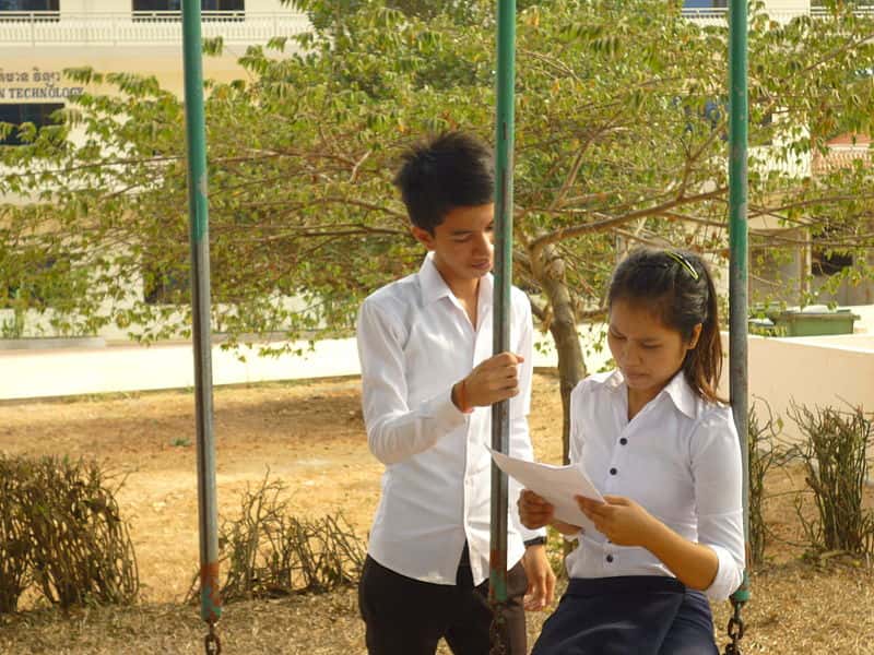Teenage_boy_and_girl,_the_girl_is_sitting_on_a_swing_reading,_the_boy_standing_next_to_her