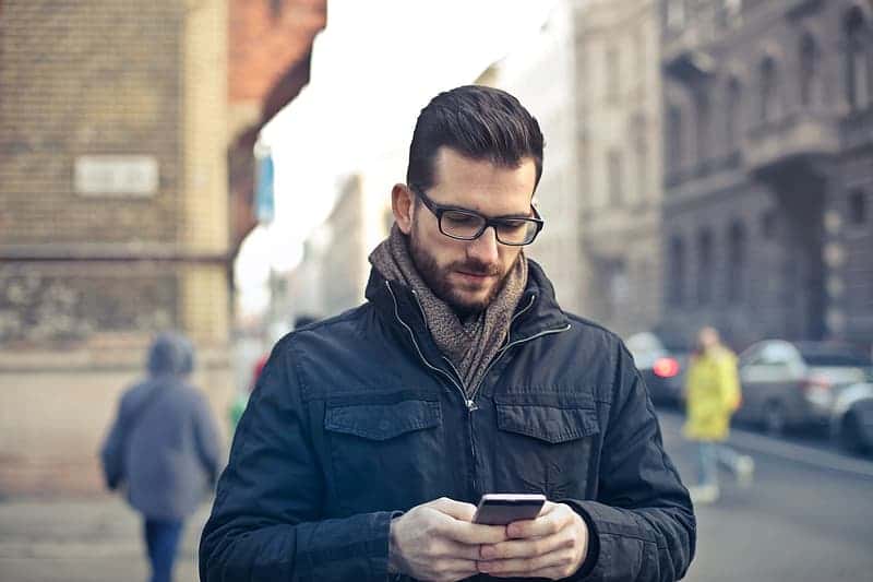 young-man-in-black-winter-jacket-and-spectacles-using-his-smartphone-while-walking-on-the-road