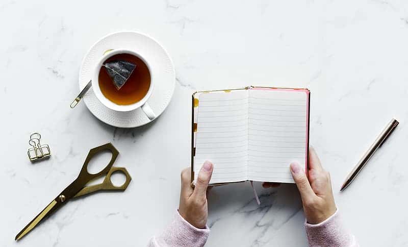 person-holding-white-book-with-black-scissors-and-white-ceramic-mug-with-coffee