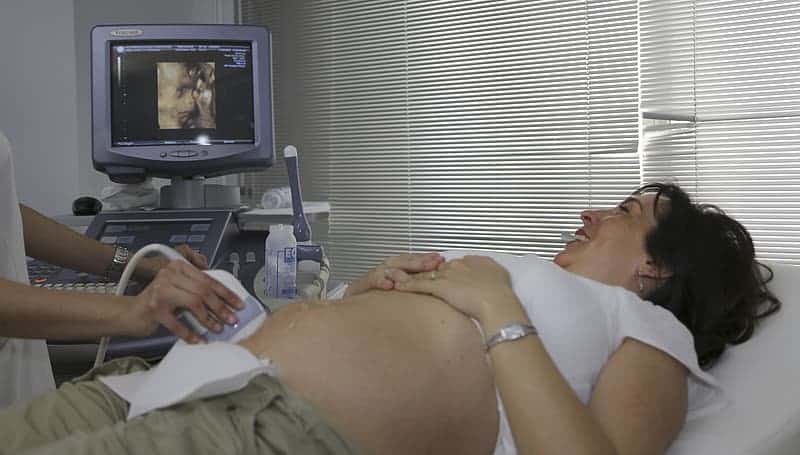 woman-undergoing-for-an-ultrasound-wearing-white-shirt-and-grey-bottoms-laying-on-white-mattress-near-grey-monitor